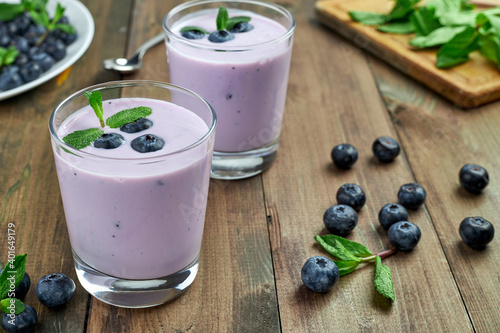 Blueberry yogurt with fresh berries and mint leaves. Two glasses of yogurt and a lot of scattered blueberries on a wooden table. Healthy breakfast.