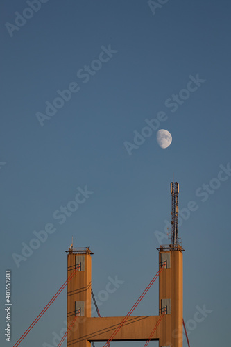 Moon shining above polyns of a suspension bridge with late afternoon sunlight