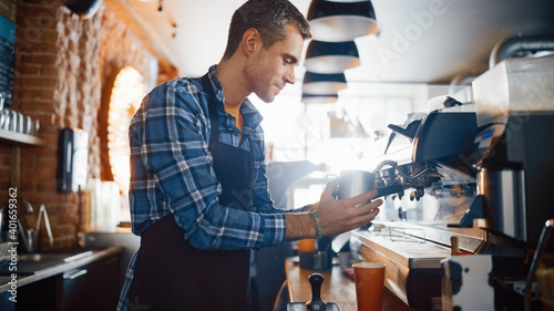 Handsome Male Barista in Checkered Shirt is Making a Take Away Cappuccino in Disposable Recycle Cup for a Customer in a Coffee Shop Bar. Female Cashier Works at a Cozy Cafe Counter in the Background.
