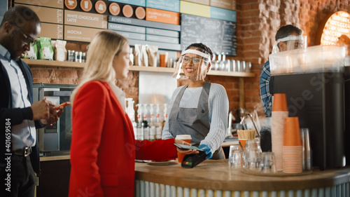 Female Customer Pays for Coffee and Pastry with Contactless NFC Payment Technology on Smartphone to a Barista in Face Shield in Cafe. Social Restrictions Concept in a Bar During Coronavirus Pandemic.