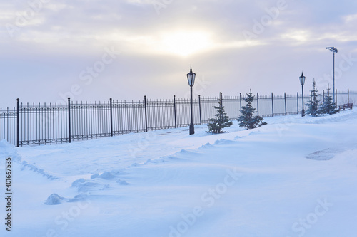 winter snowy park with a fence, lanterns and sunlight shining through the clouds