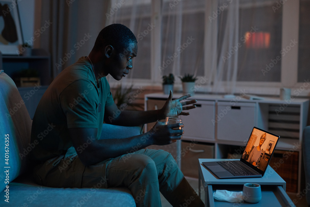African man talking to therapist online on laptop in the dark room at home
