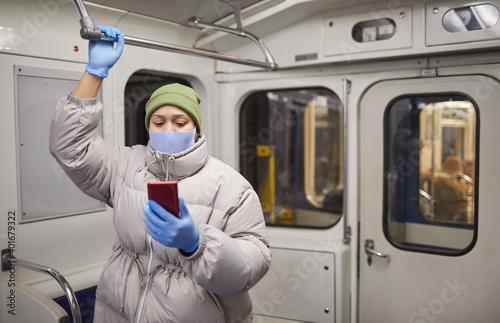 Woman in warm clothing and in protective mask and gloves reading online book on the phone while standing on a train