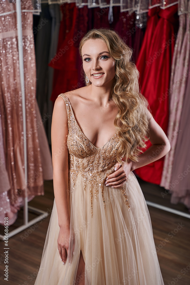 Beautiful blonde girl wearing a full-length yellow champaign chiffon slit prom ball gown decorated with golden sequins. Model in dress hire service with many dresses in different color and style.