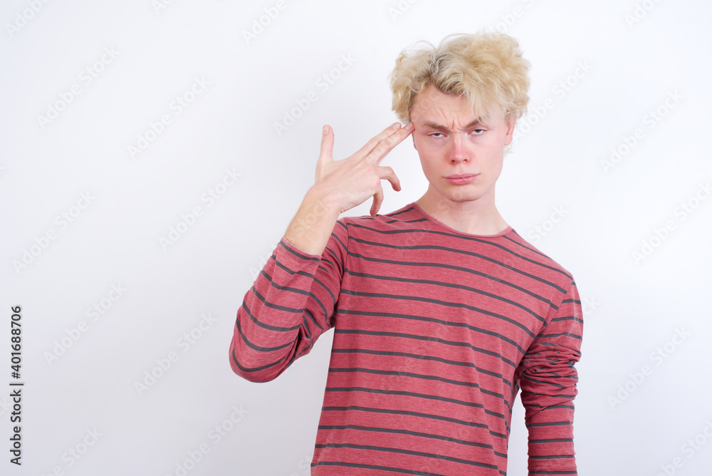 Unhappy Young handsome Caucasian blond man standing against white background makes suicide gesture and imitates gun with hand, curves lips, keeps two fingers on temple Unhappy Young handsome Caucasian blond man standing against white background makes suicide gesture and imitates gun with hand, curves lips, keeps two fingers on temple