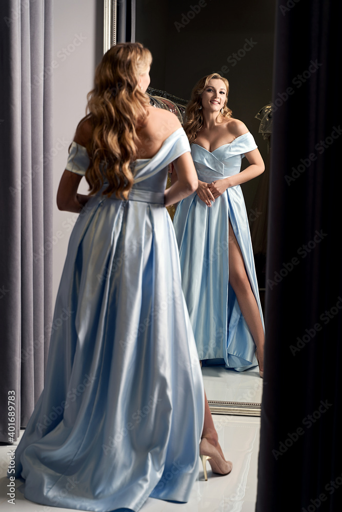 Young beautiful blonde girl wearing an off-the-shoulder full-length sky blue satin slit prom ball gown. Smiling model looking in mirror. Fitting room in dress hire service.
