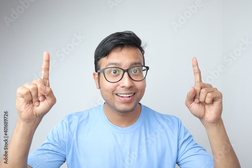 a young indian man in spectacles looking towards the camera and smiling very excited and pointing both fingers up to blank copy space