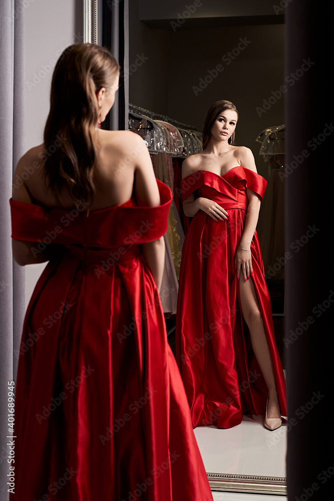 Young beautiful girl wearing an off-the-shoulder full-length crimson red satin slit prom ball gown. Model looking in mirror. Fitting room in dress hire service.