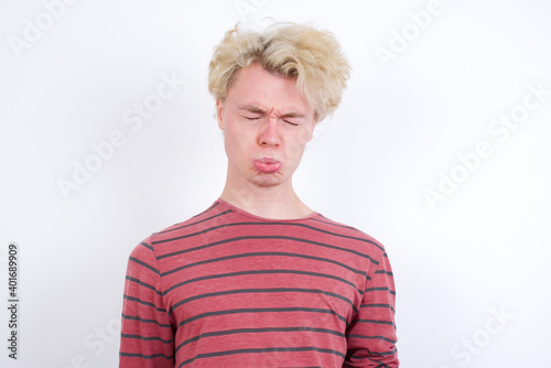 Dismal gloomy rejected Young handsome Caucasian blond man standing against white background has problems and difficulties, curves lower lip and closes eyes in despair, being in depression