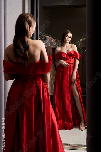 Young beautiful girl wearing an off-the-shoulder full-length crimson red satin slit prom ball gown. Model looking in mirror. Fitting room in dress hire service.