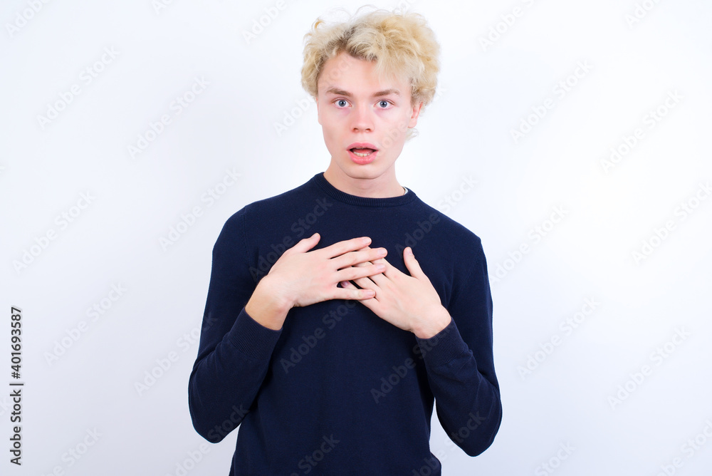 Scared Young handsome Caucasian blond man standing against white looks with frightened expression, keeps hands on chest, being puzzled to notice something strange, People, hush reaction and emotions. Scared Young handsome Caucasian blond man standing against white looks with frightened expression, keeps hands on chest, being puzzled to notice something strange, People, hush reaction and emotions.