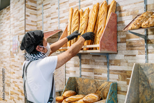selective focus of Latino baker shelving freshly baked bread with a protective face mask for the 2020 covid 19 coronavirus pandemic