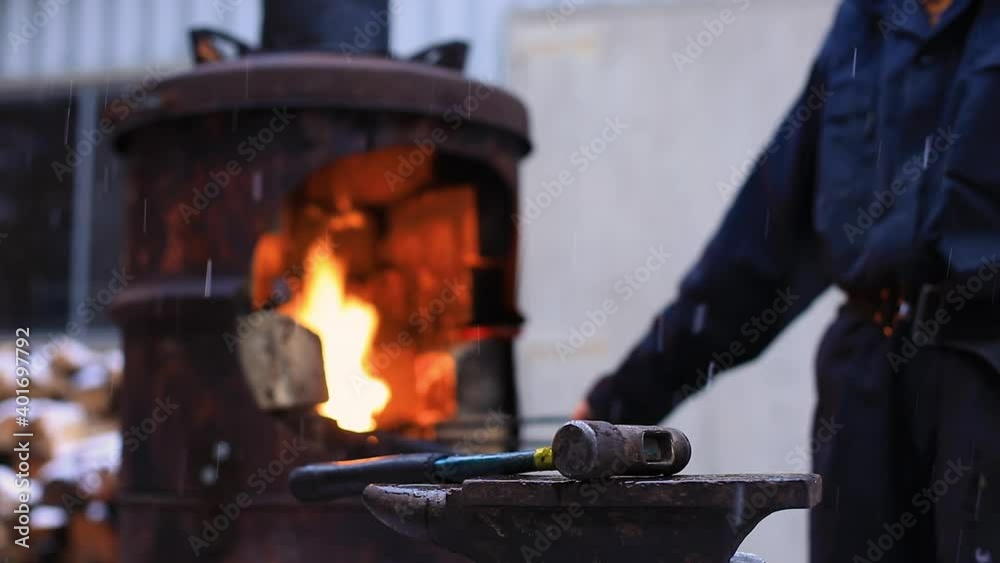 Selective focus of anvil and hammer on stool with young female craftswoman standing in front of forge wearing uniform and pocket belt Selective focus of anvil and hammer on stool with young female craftswoman standing in front of forge wearing uniform and pocket belt