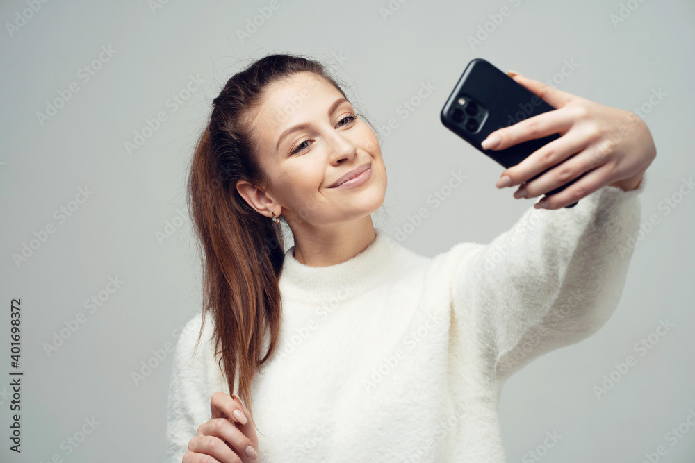holding a phone taking a selfie smiling at a picture for parents. young confident female student. Clothing white fluffy warm stylish jacket. copy space holding a phone taking a selfie smiling at a picture for parents. young confident female student. Clothing white fluffy warm stylish jacket. copy space