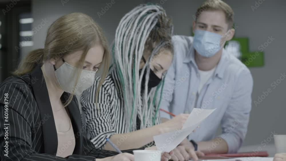 Male and female colleagues in Covid face masks discussing paperwork in office on coronavirus pandemic outbreak. Confident young Caucasian men and women talking project on Covid-19 lockdown. Male and female colleagues in Covid face masks discussing paperwork in office on coronavirus pandemic outbreak. Confident young Caucasian men and women talking project on Covid-19 lockdown.