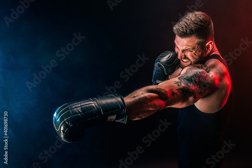 Bearded tattooed sportsman muay thai boxer in black undershirt and boxing gloves fighting on dark background with smoke. Sport concept. © zamuruev