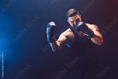 Bearded tattooed sportsman muay thai boxer in black undershirt and boxing gloves fighting on dark background with smoke. Sport concept.