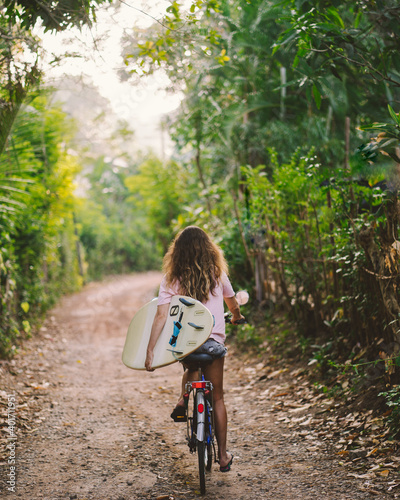 Woman on a bicycle with surfboard on rural road with green trees along. Tropical green. Sport Lifestyle concept.