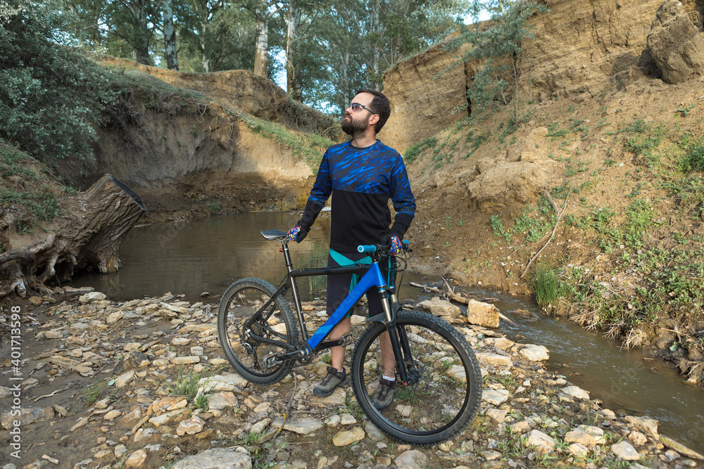 Obraz premium Cyclist in shorts and jersey on a modern carbon hardtail bike with an air suspension fork standing on a cliff against the background of fresh green spring forest
