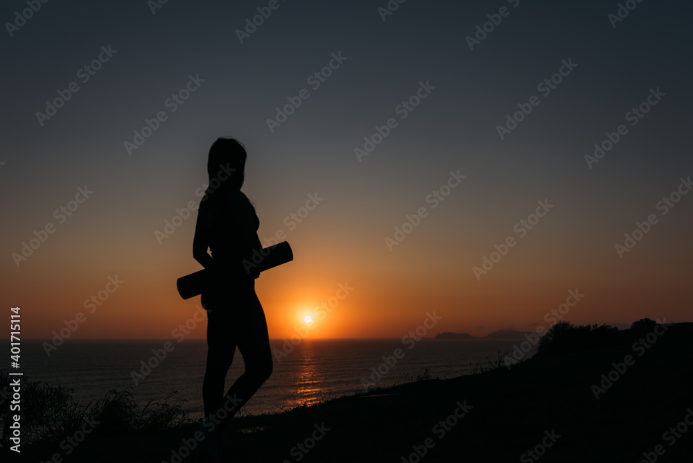 Stock image of a silhouette of a a woman looking at the sunset holding her mat after a workout yoga concept copy space