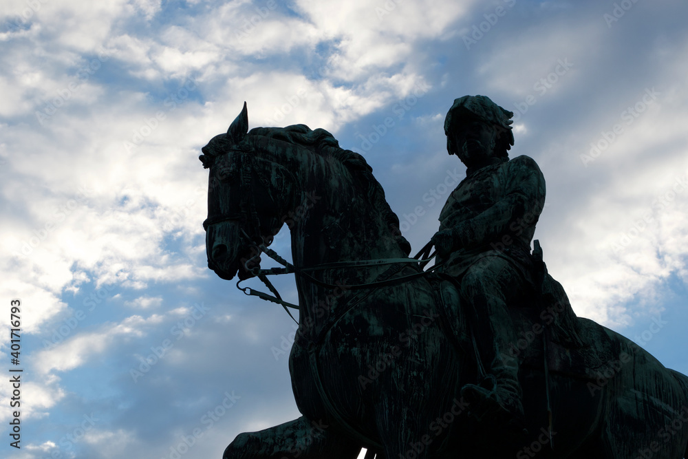 Fototapeta premium Silhouette of a dark metal statue of a soldier on a horse, standing boldly against a blue, cloudy sky.