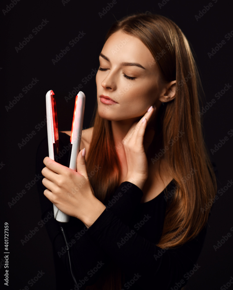 Fototapeta premium Sensual young woman with healthy silky straight hair stands with eyes closed holding white modern hair straightener in hand over dark background. Haircare, beauty, wellness concept