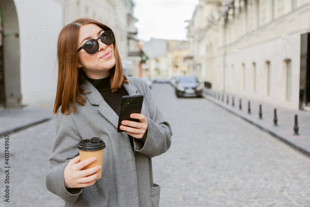 Young millennial smiling woman with smooth hair dressed in an autumn coat and sunglasses uses a smartphone and holds a cup of coffee on the go in a European city.