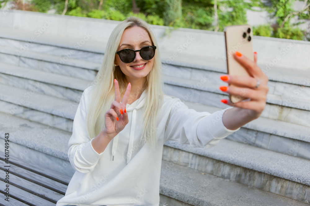 Young attractive blonde woman in sunglasses makes selfie while sitting on bench in city park