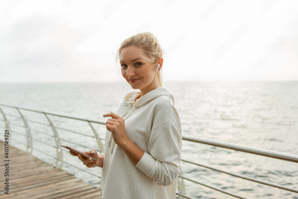 Young fitness woman blonde in a white hoodie uses a smartphone and listens to music on headphones on the beach