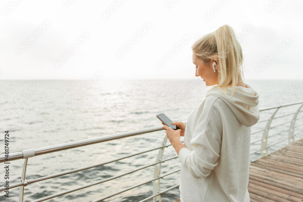 Young fitness woman blonde in a white hoodie uses a smartphone and listens to music on headphones on the beach
