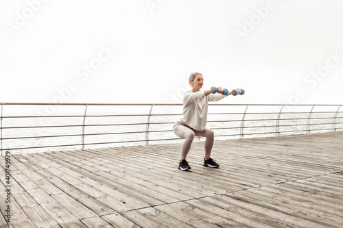 Young healthy fit woman practicing squats with dumbbells on the city beach. Healthy lifestyle