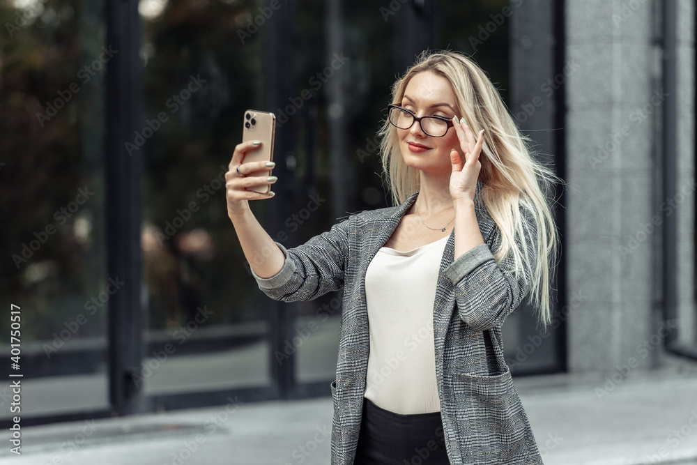Attractive blonde business woman makes selfie on a smartphone on the background of a business office.
