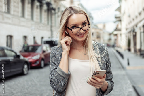 Portrait of confident business woman in urban street. Blond girl in business suit using smartphone and listening to music with wireless headphones