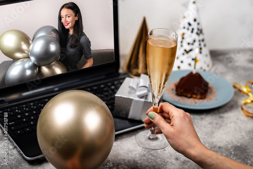 Birthday party in online video call. Female hands are holding glass of wine and balloon in front of laptop screen. Young woman celebrating her birthday at home. Remote Celebration Concept.