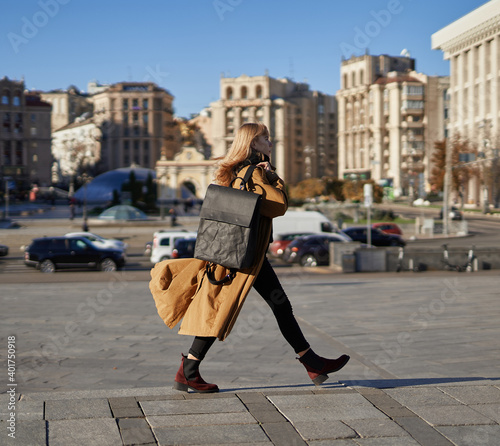 Foto Attractive blonde woman in orange trench walking in a hurry with recycled made paper backpack on blue sky background of the european city streets