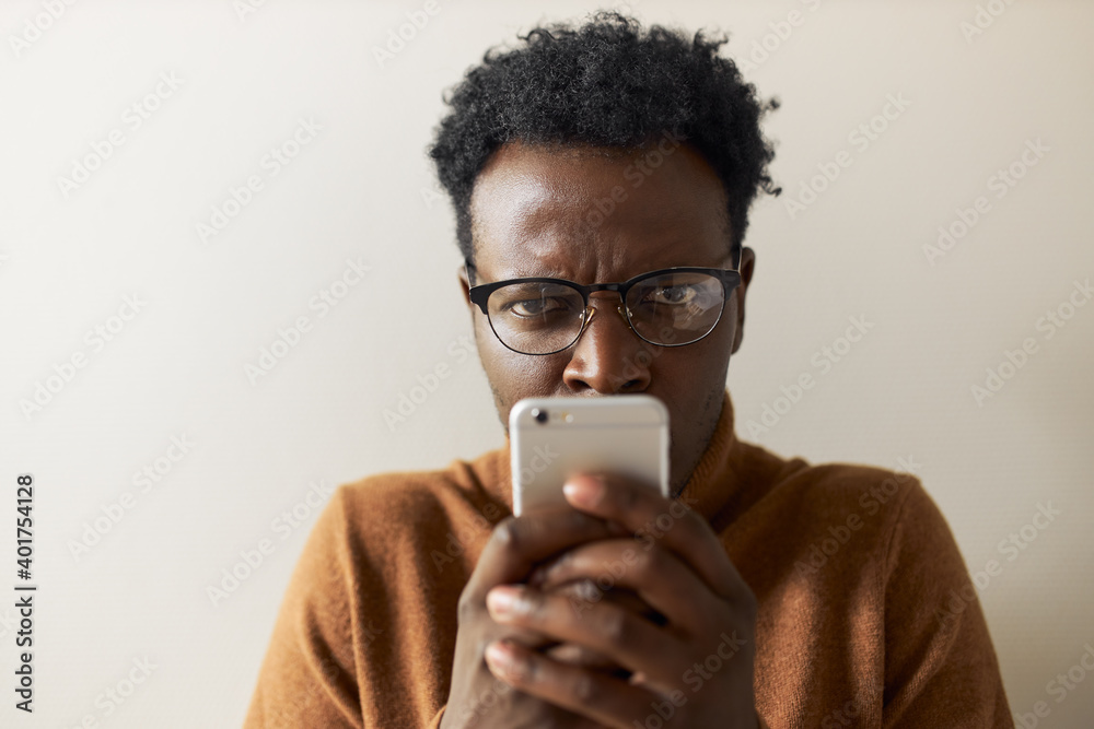 Distrustful frowning young dark skinned man posing isolated with smart phone at his face, having suspicious facial expression, spying on his girlfriend, checking messages in social networks