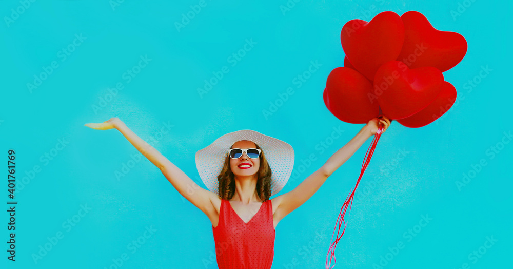 Beautiful happy young woman raising hands up with bunch of red heart shaped balloons wearing a summer straw hat and sunglasses on a blue background