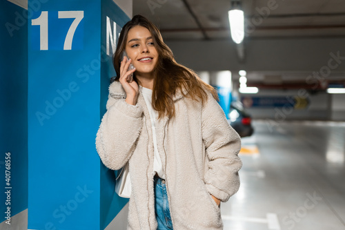 Stylish smiling woman in the underground parking talking on the phone leaning against the column