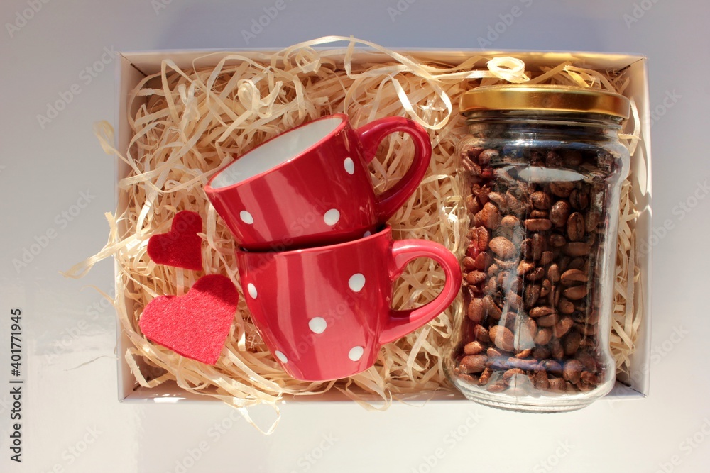 Valentine's day gift box with two red espresso coffee cups, coffee beans in glass jar, and red hearts. Flat lat composition