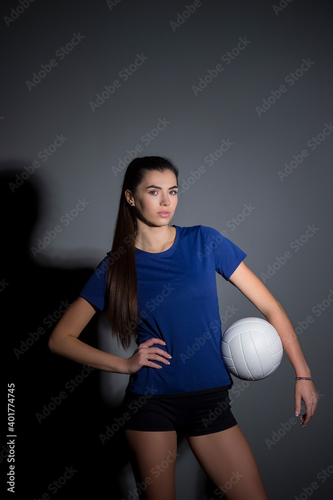 Volleyball girl hold and kick ball in blue costume on dark wall background. Player doing sport workout at home. Sport and recreation concept Volleyball girl hold and kick ball in blue costume on dark wall background. Player doing sport workout at home. Sport and recreation concept