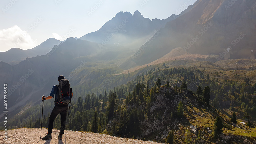 A man with a big hiking backpack standing at the edge of a mountain and admiring the panoramic view in front of him. Sharp and high mountains around. Dense forest at the foothill. Freedom and peace