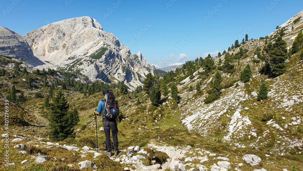 A man with a big hiking backpack hiking in Italian Dolomites. He walks on a very narrow pathway. There are high and stony mountain chains in front. The slopes are overgrown with a few trees. Solitude