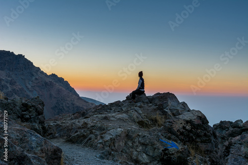 A female tourist sitting on the peak of a rocky mountain watching sun rising in Darband valley in autumn in dawn against colorful sky in the Tochal mountain.