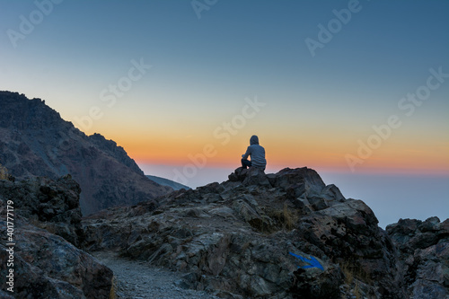 A tourist sitting on the peak of a rocky mountain watching sun rising in Darband valley in autumn in dawn against colorful sky in the Tochal mountain.