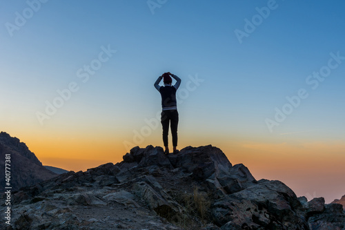 A female tourist standing on the peak of a rocky mountain watching sun rising in Darband valley in autumn in dawn against colorful sky in the Tochal mountain.