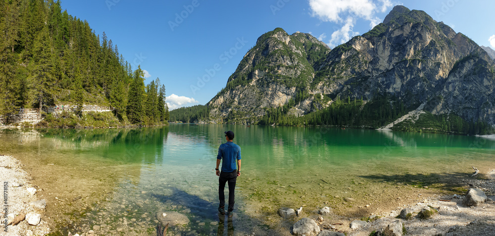 A man standing on a boulder at the shore of the Pragser Wildsee, a lake in South Tyrolean Dolomites. High mountain chains around the lake. The sky and mountains are reflecting in the lake. Calmness A man standing on a boulder at the shore of the Pragser Wildsee, a lake in South Tyrolean Dolomites. High mountain chains around the lake. The sky and mountains are reflecting in the lake. Calmness