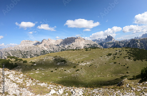 A panoramic view on a vast valley in Italian Dolomites. There are high mountain chains around. The bottom of the valley is lush green. A few trees on the slopes. Few soft clouds on the sky. Freedom photo