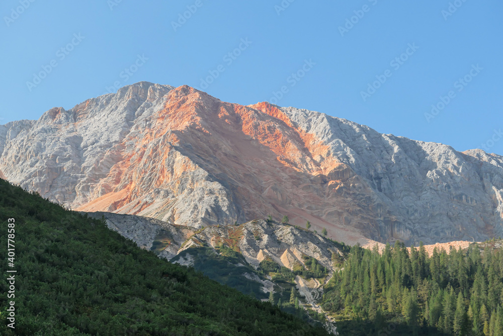 A close up view on a distant mountain peak in Italian Dolomites. The mountain has distinctive red color, mixing with grey sides. At the foothill there is a dense forest. Discovering the nature