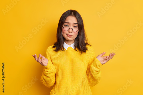 Unaware hesitant Asian woman shrugs shoulders in clueless gesture cannot make decision wears round spectacles and casual jumper isolated over yellow background. Indifferent cluess female model photo