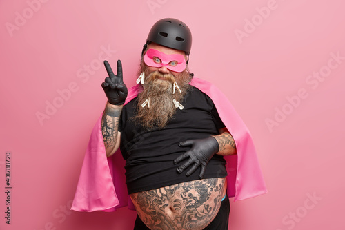 Portrait of funny bearded superhero makes peace gesture has big tattooed belly wears mask helmet and cape isolated over pink background. European adult man shows victory sign being on costume party